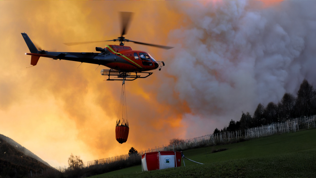 Hubschrauber mit rotem Löschbehälter fliegt vor orangefarbenem Rauch über grünem Feld mit Feuerwehrzelt.