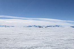 Eine weite, schneebedeckte Landschaft unter blauem Himmel. Im Hintergrund sind schneebedeckte Berge zu sehen. Der gesamte Bereich wirkt kalt und unberührt, mit wenigen Spuren menschlicher Aktivität.