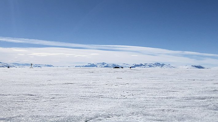 A vast, snowy landscape stretches toward the horizon under a clear blue sky. Snow-covered mountains are visible in the background. The bright, reflective surface of the snow enhances the sense of openness in this arctic environment.