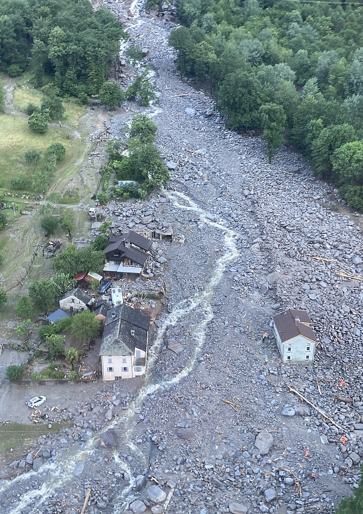 Luftaufnahme eines Murgangs mit einem von Geröll und Wasser durchzogenen Flussbett, umgeben von mehreren Häusern und Bäumen.