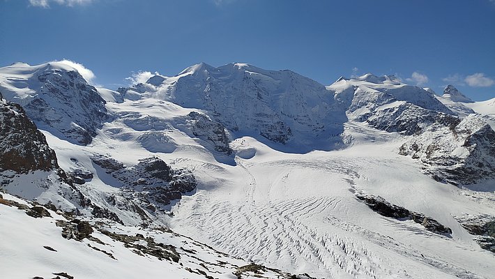 Vue majestueuse de montagnes enneigées sous un ciel bleu clair. Les glaciers descendent des sommets, créant des crevasses et des lignes de glace. Le paysage est immaculé, avec des pentes douces couvertes de neige brillante.