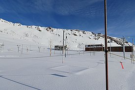 A snowy landscape features a research station and various measurement equipment poles spread across the foreground. Snow covers the ground, while a rocky mountain rises in the background under a clear blue sky. The scene illustrates an environment for scientific studies in a remote location.
