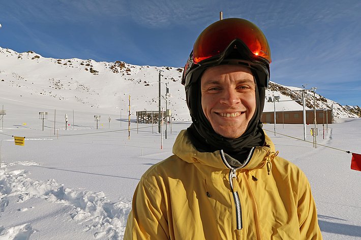 A smiling young man wearing a yellow jacket and ski helmet stands in a snowy landscape, with a mountain in the background. The scene suggests a ski resort, featuring clear skies and snow-covered terrain. Snow trails and ski lift poles are visible behind him.