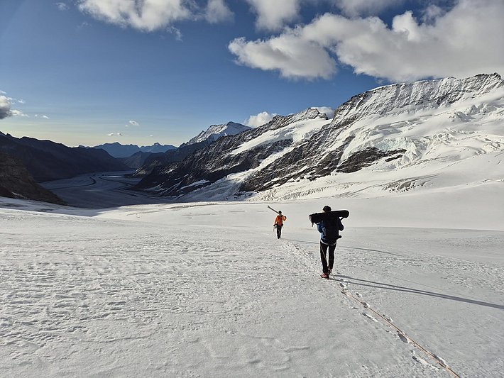 Two people carrying ski equipment walk across a vast snow-covered glacier surrounded by snow-capped mountains under a blue sky with clouds.
