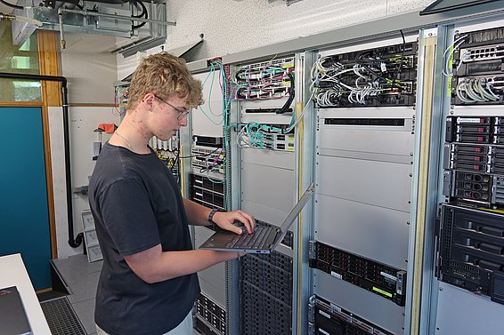 Un jeune homme se tient devant une baie de serveurs, utilisant un ordinateur portable. Des câbles sont visibles derrière lui, connectés aux équipements informatiques. L'environnement semble être une salle de serveurs ou un centre de données.