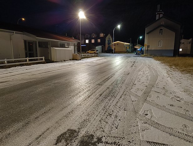 A nighttime scene of a quiet, snowy street. Streetlights illuminate the road, which shows tracks from vehicles. On the right, there is a church and several buildings, while the grassy area on the side contrasts with the white snow covering the road.