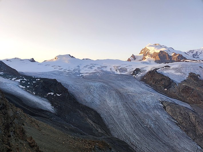 Glacier with visible ice and rock surfaces, mountains in the background at sunrise or sunset.