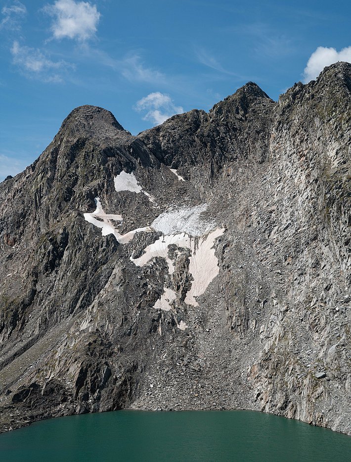 Green mountain lake at the base of steep rocky cliffs with patches of snow under a blue sky.