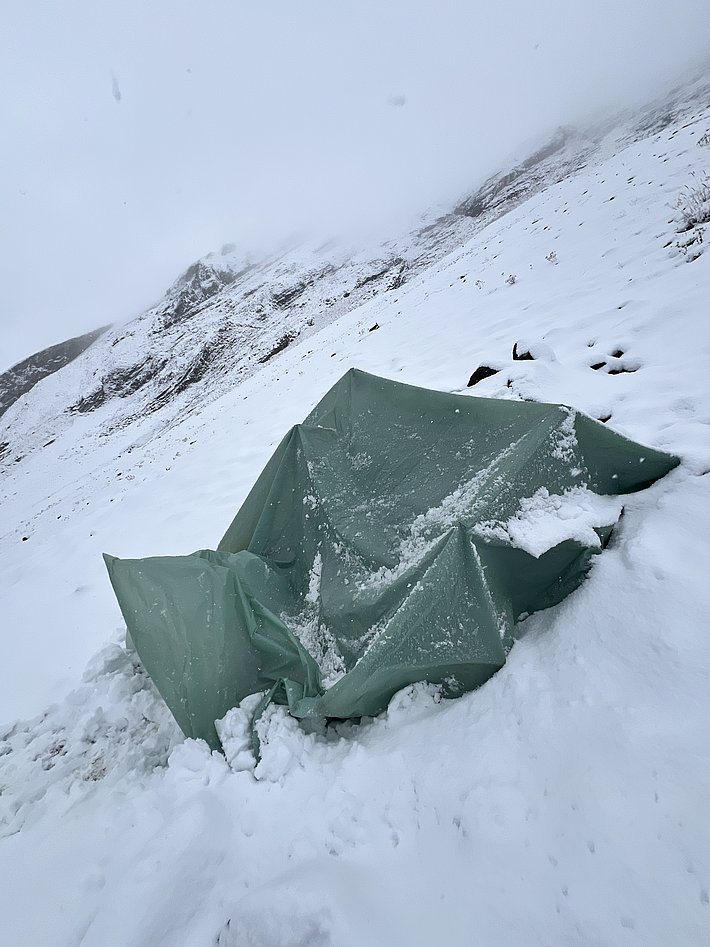 A green tent partially covered in snow, set against a backdrop of a snowy landscape with mountains. The scene is overcast, indicating cold weather conditions.