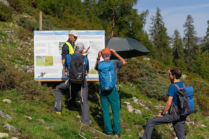 Vier Personen stehen vor einem Informationsschild im Freien. Eine Person trägt eine gelbe Weste, die anderen einen Rucksack. Im Hintergrund sind Bäume und Sträucher sichtbar.