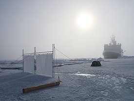 Eine Forschungsstation auf dem Eis mit einer weißen Leinwand, einem grünfarbenen Zelt und einem Schiff im Hintergrund. Die Sonne steht hoch am Himmel.