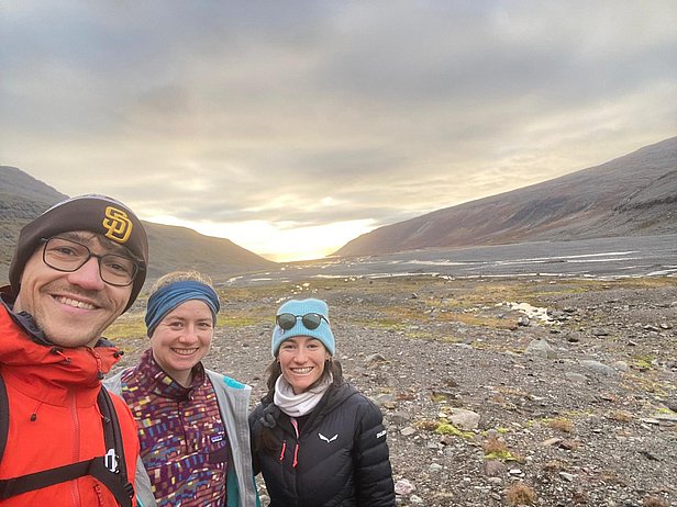 Three hikers pose for a selfie outdoors, with mountains and a valley in the background. The group is smiling, dressed in outdoor gear, and the light of a sunset is visible, adding warmth to the scene.