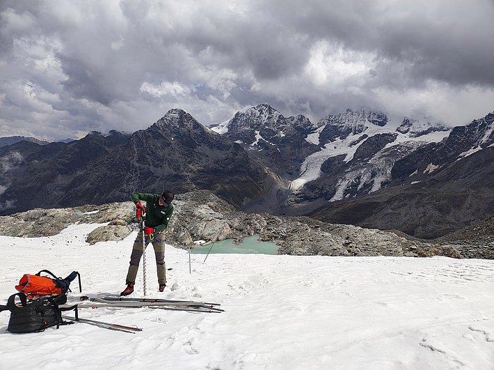 Person in green jacket using a red eisbohrer on a snowy mountain plateau with rocky peaks and cloudy sky.