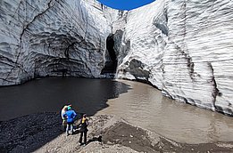 Three people stand on the shore of a meltwater lake in front of tall, layered glacier walls with a dark cave in the center under a blue sky.