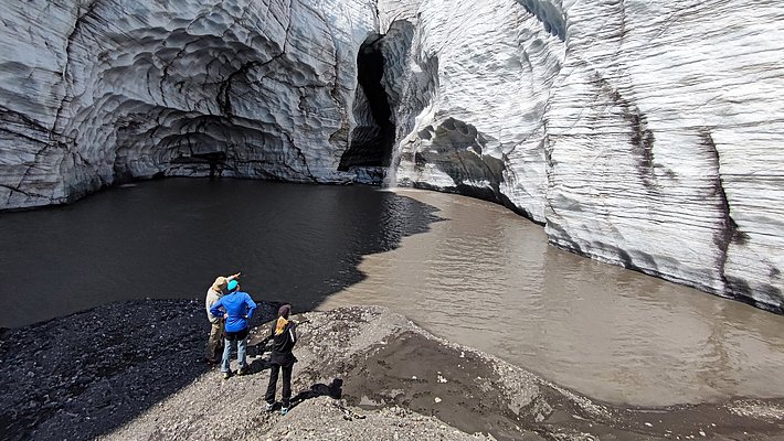 Three people stand on the shore of a meltwater lake in front of tall, layered glacier walls with a dark cave in the center under a blue sky.