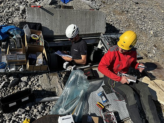 Two individuals are working outdoors near an open manhole. One person, wearing a white helmet, is sitting and reading a document, while the other, in a yellow helmet, is assembling equipment. Various tools and cables are spread out on a rocky surface around them.