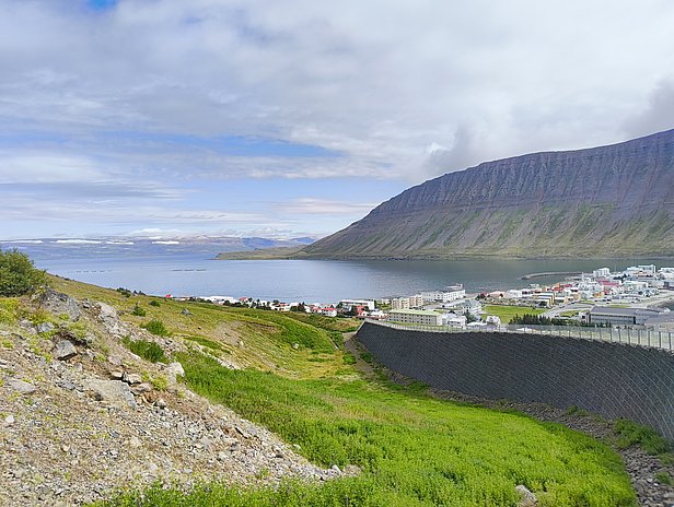 A panoramic view of a coastal town nestled beside a calm bay, surrounded by lush greenery and a steep mountain range in the background. The sky is partially cloudy, adding depth to the serene landscape.