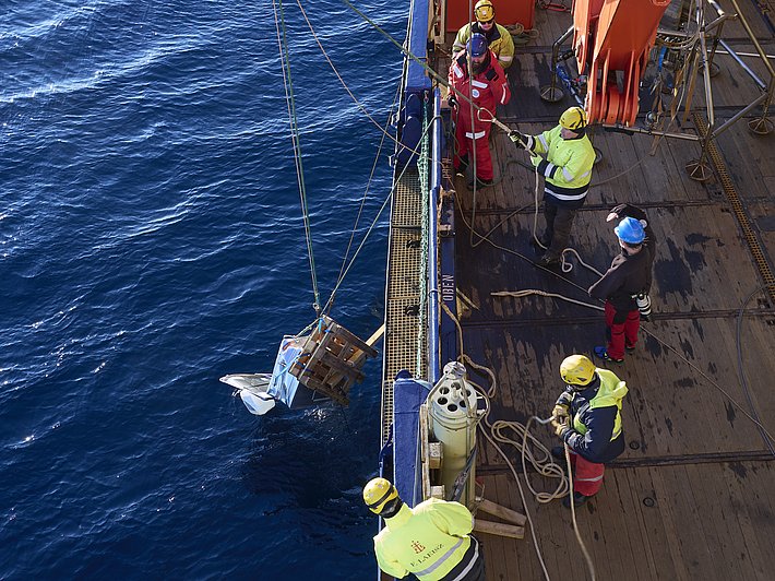 Sechs Personen in gelben und roten Arbeitskleidung auf einem Schiff, die eine große Messinstallation aus dem Wasser heben. Der Ozean ist blau.