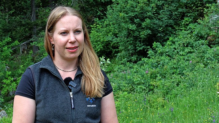 Donna con capelli biondi lunghi e maglietta nera davanti a una foresta fitta