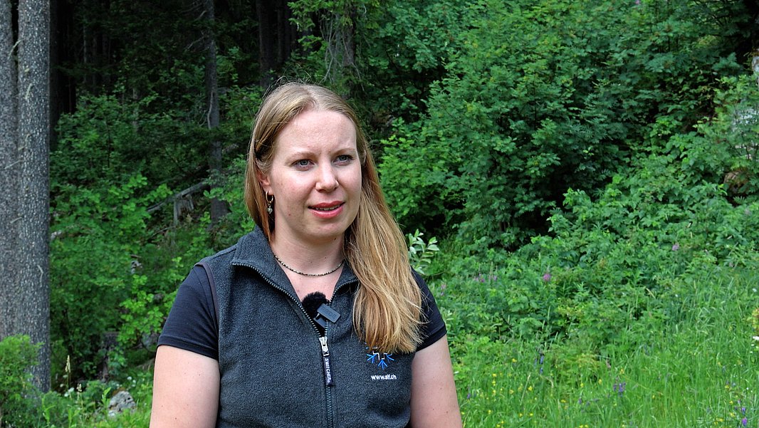 Frau mit langen blonden Haaren und schwarzem T-Shirt vor dichtem Wald