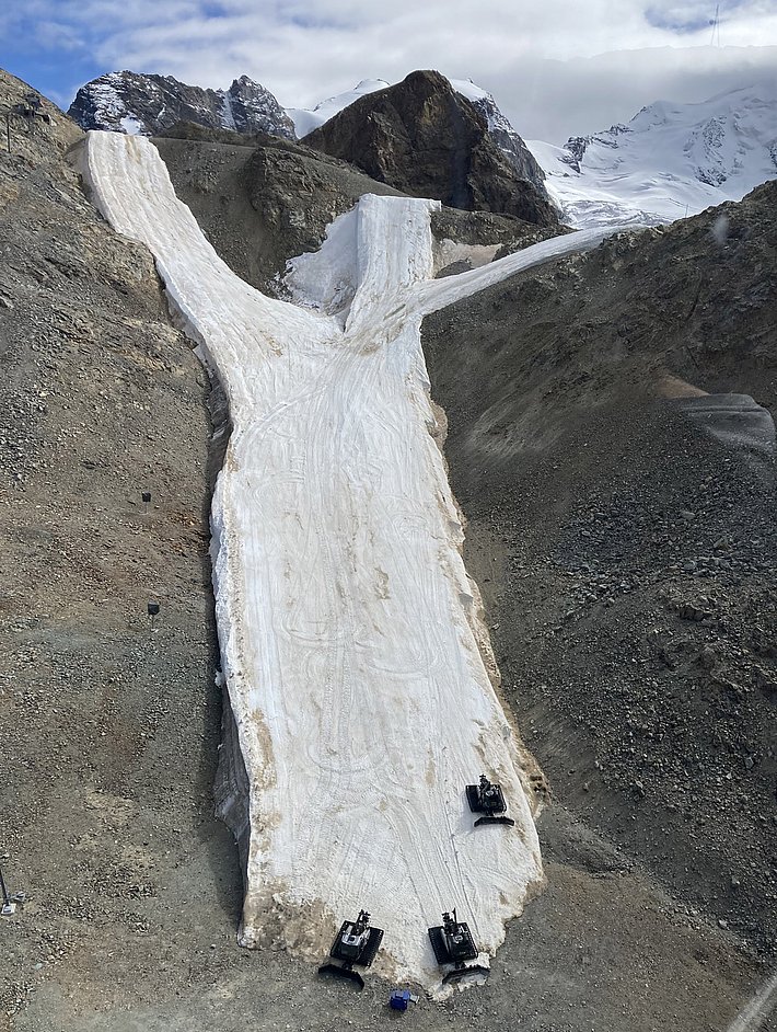 Snow-covered ski slope with three small snow grooming machines, surrounded by rocky, snow-free terrain and mountains in the background.