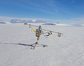 Eine Messstation steht auf einer schneebedeckten Eisfläche. Im Hintergrund sind schneebedeckte Berge und der klare Himmel sichtbar. Die Station ist mit verschiedenen Geräten ausgestattet, die zur Datenerhebung in dieser arktischen Umgebung dienen.