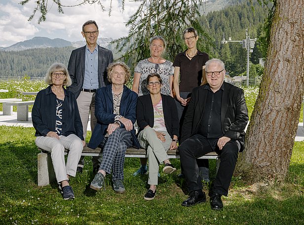 Eine Gruppe von sieben Personen sitzt auf einer Bank im Freien, umgeben von Bäumen und einer Landschaft mit Bergen im Hintergrund. Es sind sowohl Frauen als auch Männer unterschiedlichen Alters zu sehen, die freundlich in die Kamera schauen.