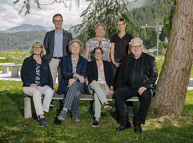 A group of seven people is seated on a bench in a scenic outdoor setting. They are positioned in front of a backdrop of greenery and mountains, with some trees providing shade. The group appears to be engaged in a professional gathering.
