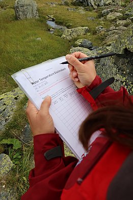 A woman in a red jacket holds a clipboard chart and pen, with rocks and grass in the background.