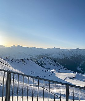 Blick auf schneebedeckte Berge bei Sonnenaufgang mit klarem Himmel, im Vordergrund ein Metallgeländer