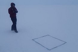 Person in roter Jacke und dunkler Hose steht auf verschneitem Boden neben einem quadratischen Rahmen und fotografiert den Schnee.