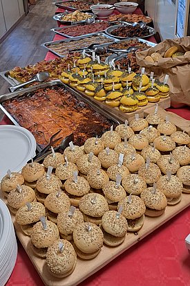 A long table is filled with various dishes, including trays of baked goods, pastries, and savory items. In the foreground, there are numerous round, topped bread rolls. 