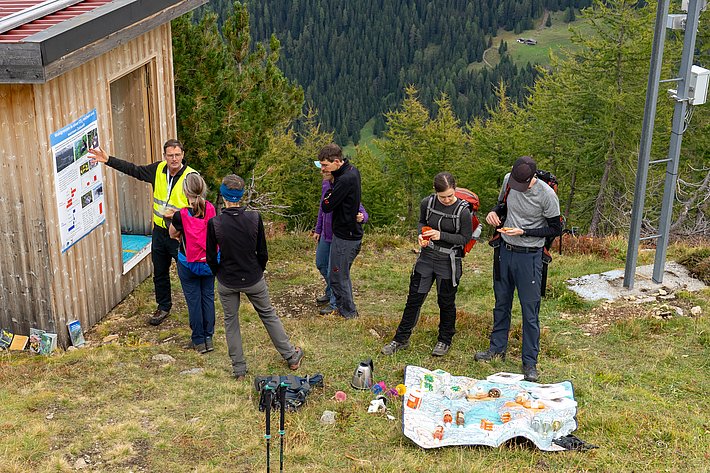 Eine Personengruppe steht vor einer Holzhütte auf einem Berg. Eine Person trägt eine gelbe Weste, während die anderen in Freizeitkleidung sind. Auf dem Boden liegt eine Karte und einige Snacks.