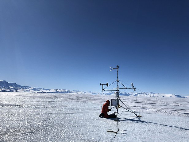 A person wearing a red jacket is kneeling next to a measuring device on the Nansen ice cap, with a breathtaking view of the snow-capped mountains and sea ice.