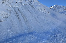 Snow-covered mountain with visible avalanche tracks under a clear blue sky, blurred propeller in the foreground.