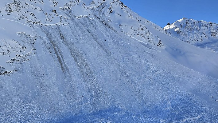 Snow-covered mountain with visible avalanche tracks under a clear blue sky, blurred propeller in the foreground.