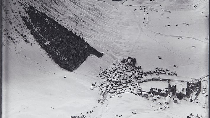 Black and white aerial photograph of a snow-covered mountain village with forest on the hillside.