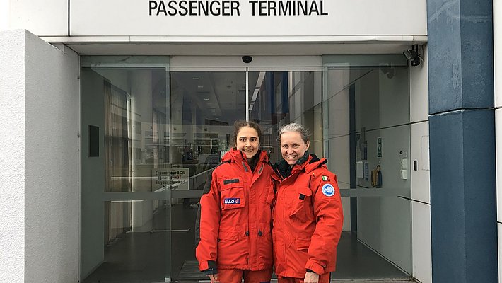 Two people in orange expedition suits stand in front of the entrance to the United States Antarctic Programme Passenger Terminal in Christchurch.
