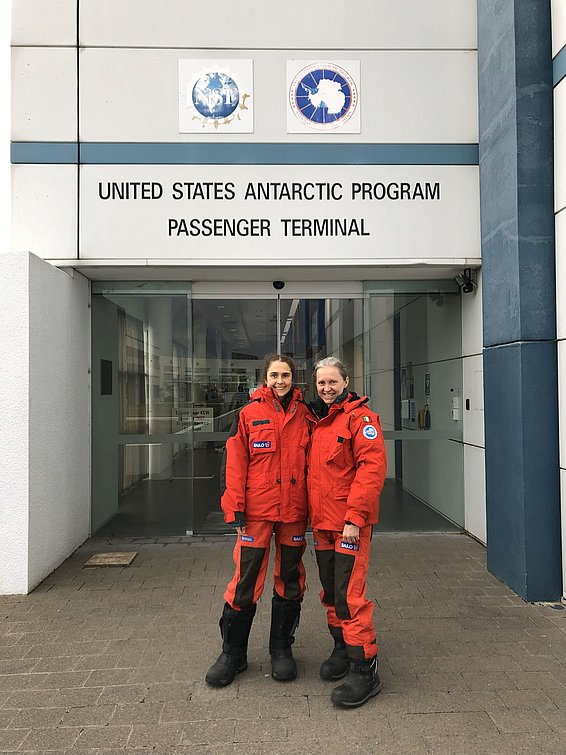 Two people in orange expedition suits stand in front of the entrance to the United States Antarctic Programme Passenger Terminal in Christchurch.