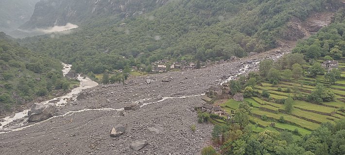 Luftaufnahme eines felsigen Erdrutschgebiets mit einem gewundenen Fluss, üppig grünen Hügeln und vereinzelten Häusern inmitten der Trümmer.