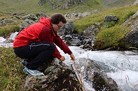A woman in a red jacket and dark trousers crouches by a mountain stream, holding a folding rule in the water.