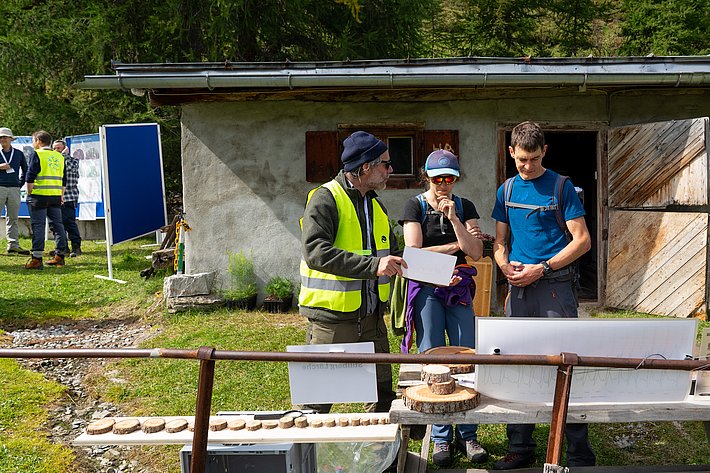 Drei Personen stehen vor einem Tisch mit Holzscheiben und einem Dokument. Im Hintergrund sind ein alter Stall und weitere Personen zu sehen.