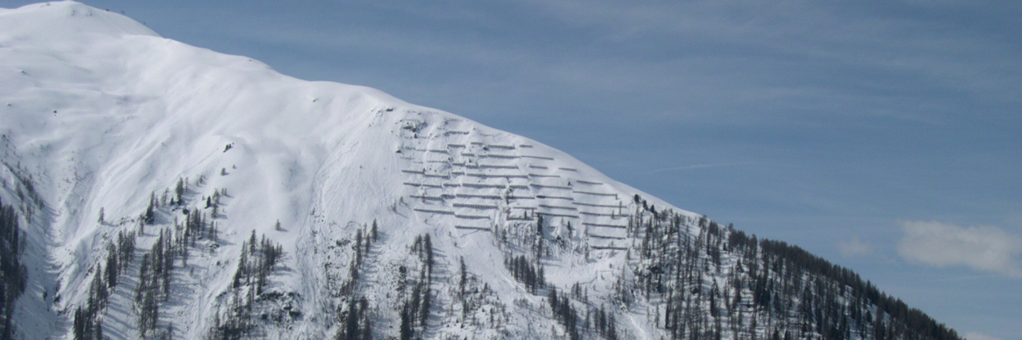 Schneebedeckter Berg mit Schutzwaldstrukturen zur Lawinenprävention unter blauem Himmel